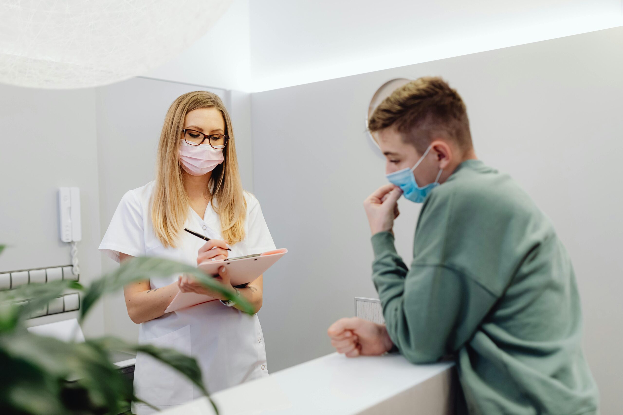 A healthcare professional in a mask discussing medical forms with a young patient at the reception desk