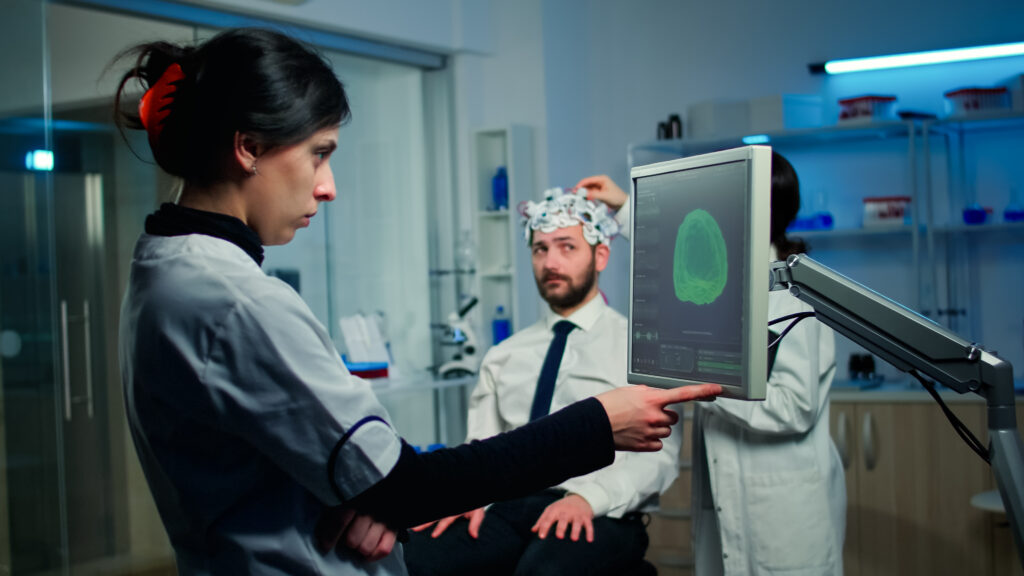 Researcher analyzing brain scan results on a monitor while a colleague conducts a test on a patient in the background